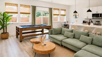 Een moderne woonkamer met een pooltafel, een groene bank en een eethoek met een witte tafel en stoelen.