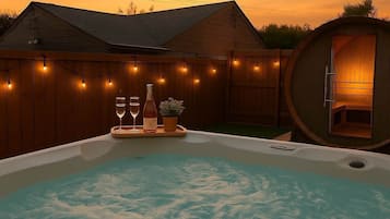 Een hottub met een houten terras en lichtjes, een houten blokhut en een houten omheining.