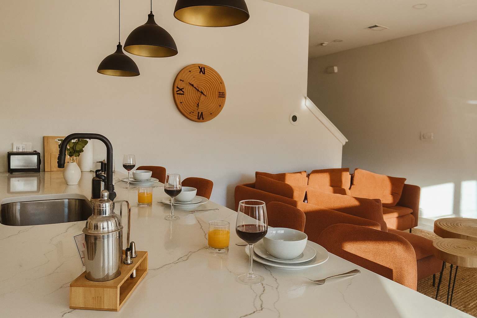 Eine moderne Küche mit Marmorarbeitsplatte, integriertem Spülbecken und einer Kaffeemaschine. An der Wand hängt eine Holz-Uhr und im angrenzenden Wohnbereich steht eine Couch.