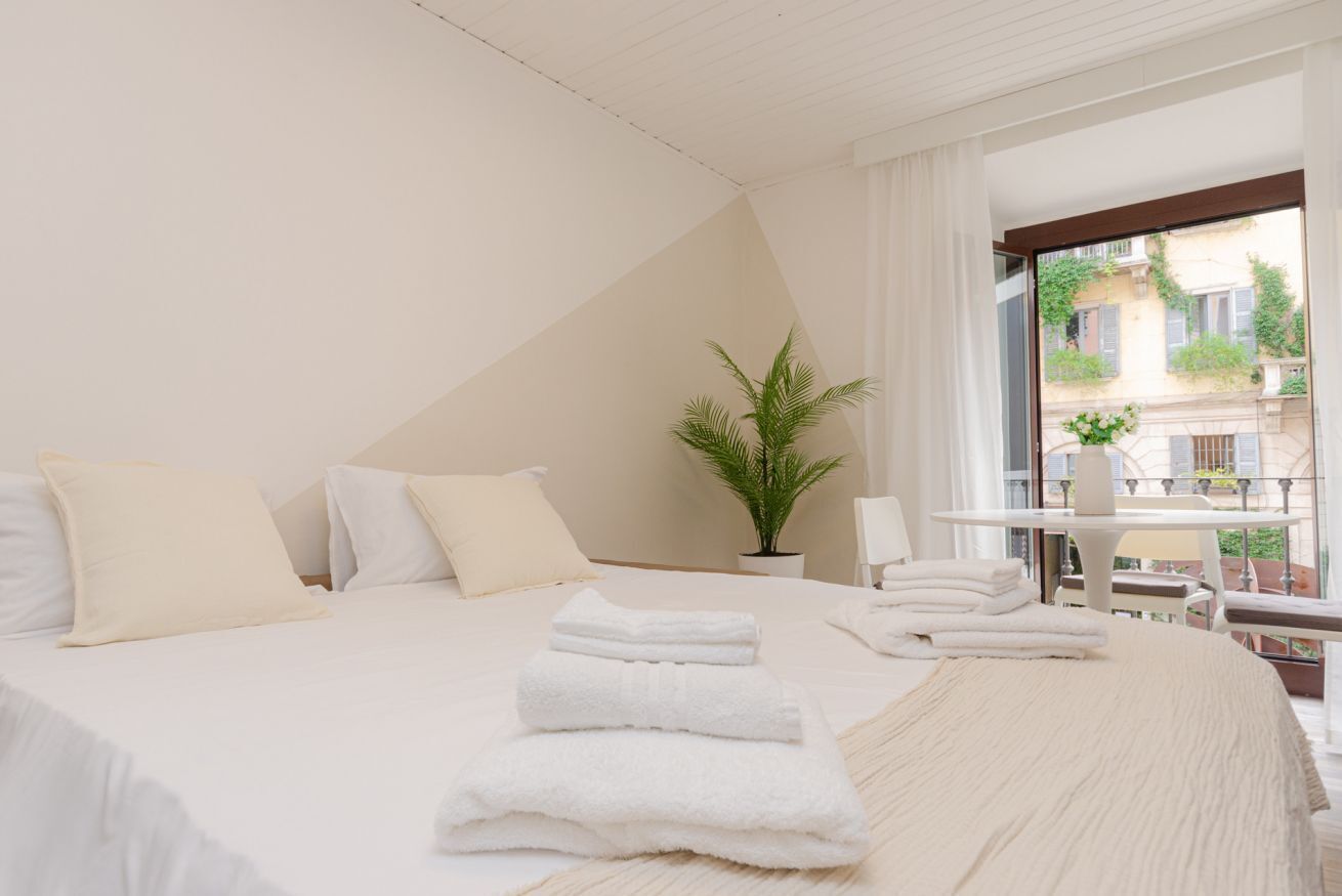A modern hotel room with a large bed, white linens, a potted plant, and a balcony with a table and chairs.