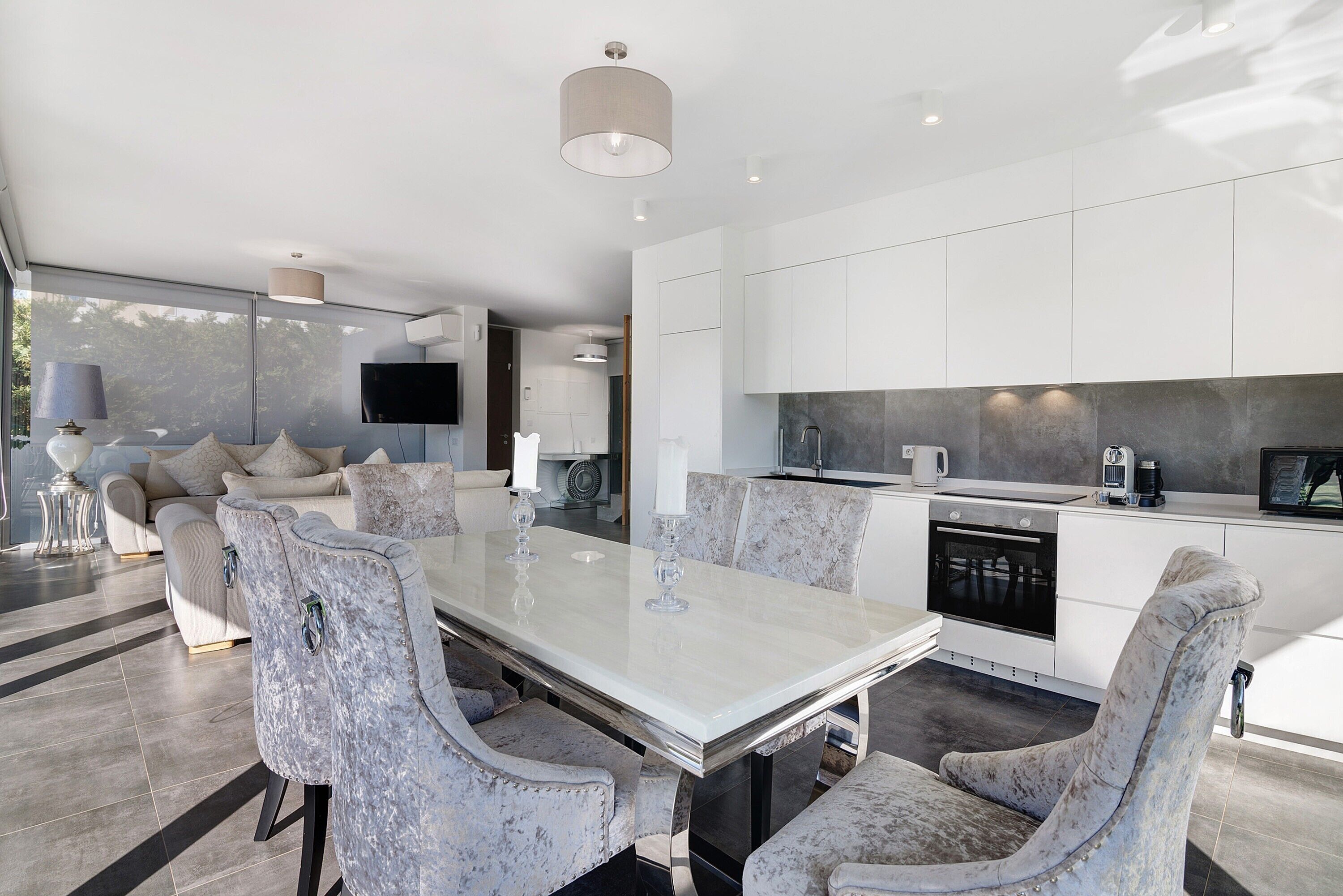 A modern kitchen with white cabinetry, a dining area with a marble table, and a living room with a sofa and TV.