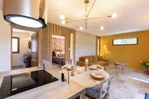 A modern kitchen with an induction cooktop, a dining area with a round table, and a living room with a sofa and a window. - Mas9Romanin:Gîte Evasion in Provence (Saint-Rémy-de-Provence)