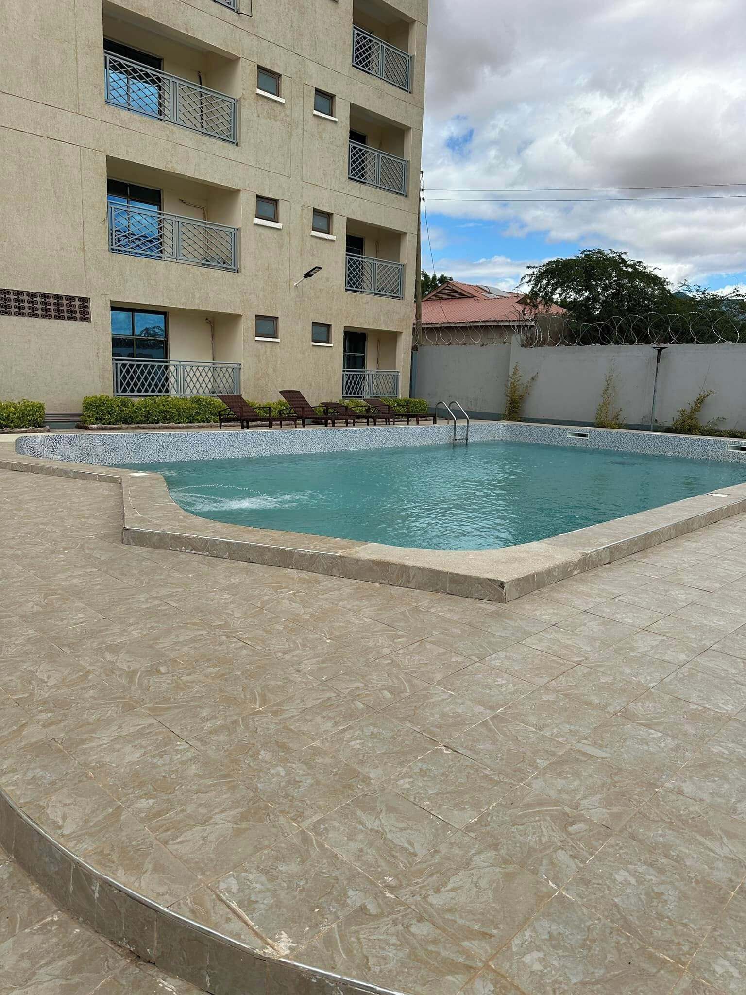A swimming pool with a tiled deck, a building with balconies, and a clear sky.
