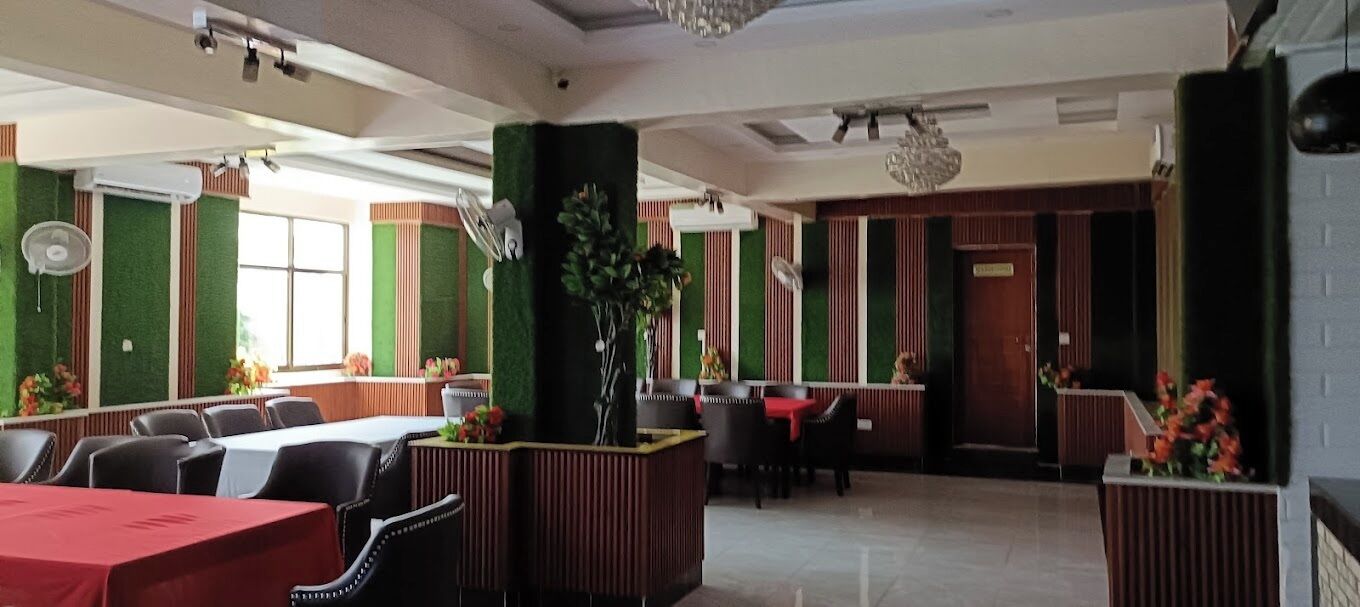 A restaurant with a modern interior, featuring green walls, wooden accents, and a central plant. Tables and chairs are arranged for dining.