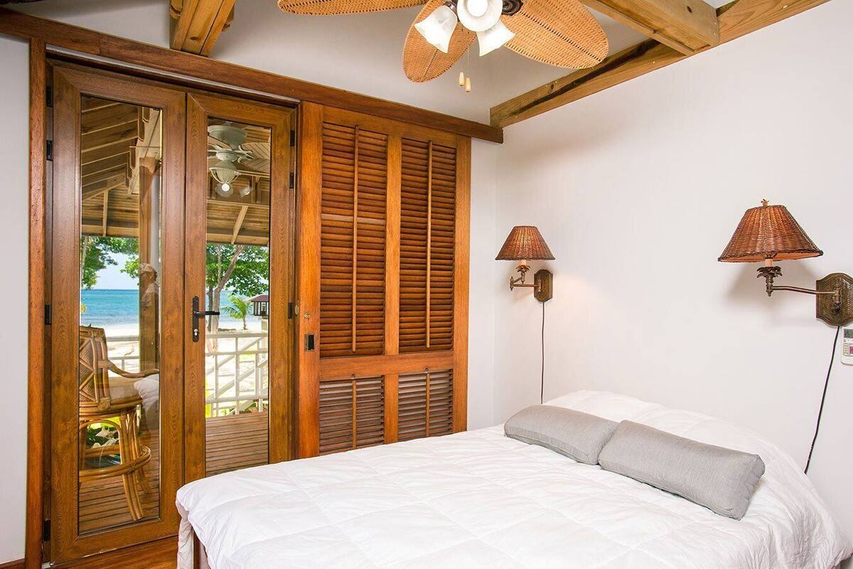 Ein Schlafzimmer mit Bett, Holzklappläden, Deckenventilator und Blick auf den Strand durch eine Glastür.