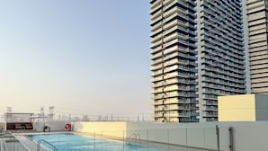 A rooftop swimming pool with a glass railing, a lounge area with chairs, and a high-rise building in the background. - PVH - Cozy 1BR - Binghatti House (Dubai)