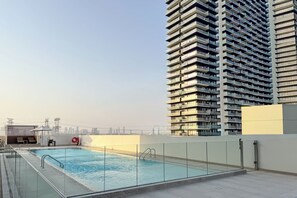 A rooftop swimming pool with a glass railing, a lounge area with chairs, and a high-rise building in the background. - PVH - Cozy 1BR - Binghatti House (Dubai)