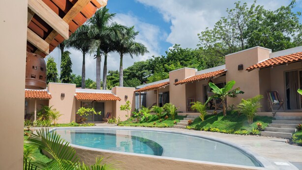 A pool area with a curved edge, surrounded by palm trees and a building with a tiled roof. - Casa Nära Hotel (San Juan del Sur)