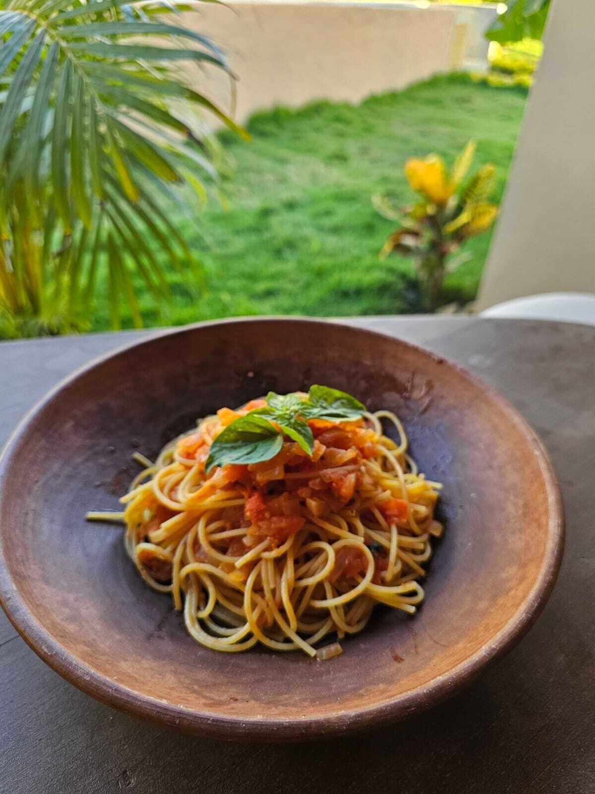 Eine Schüssel Spaghetti mit Tomatensauce und Basilikoblatt, auf einer dunklen Oberfläche, unscharfer Hintergrund draußen.