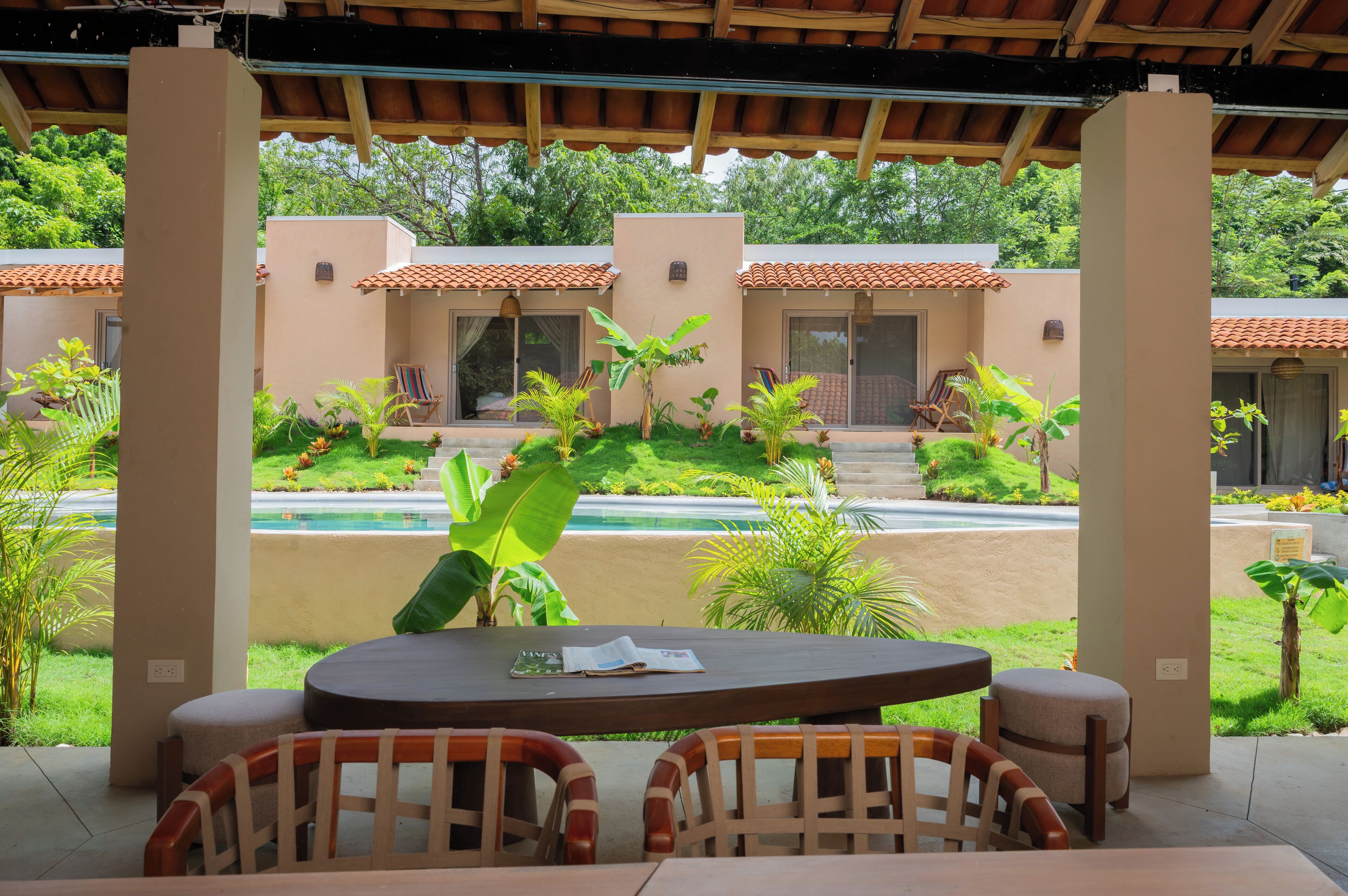 A covered outdoor seating area with a table and chairs, overlooking a pool and a building with a tiled roof.