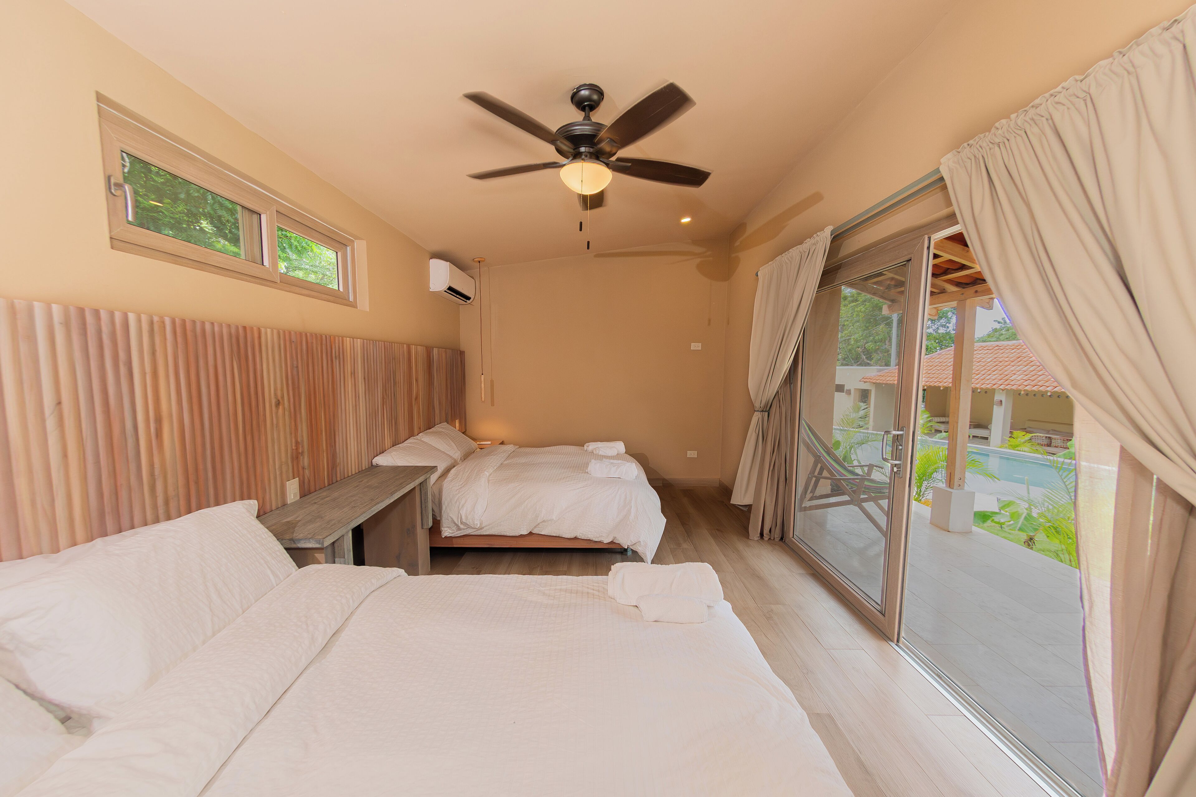 A hotel room with two beds, a ceiling fan, a window with curtains, and a view of a pool and greenery outside.