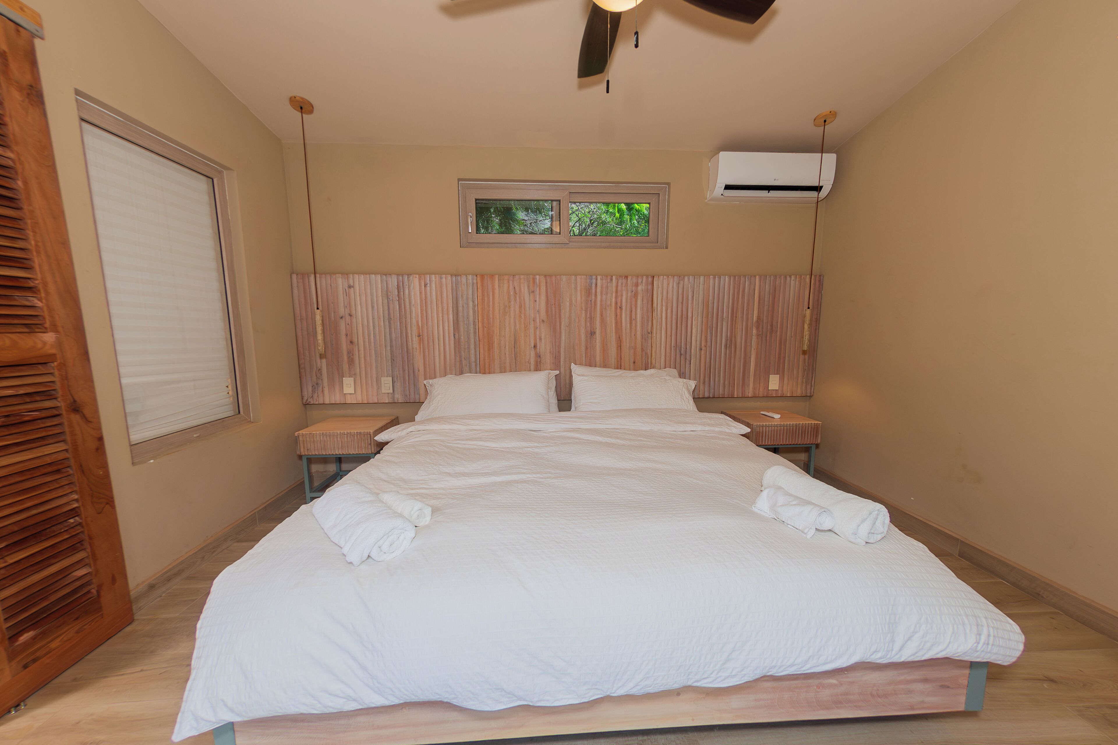 Een slaapkamer met een groot bed, een houten hoofdbord, twee nachtkastjes, een raam met uitzicht en een airconditioning.