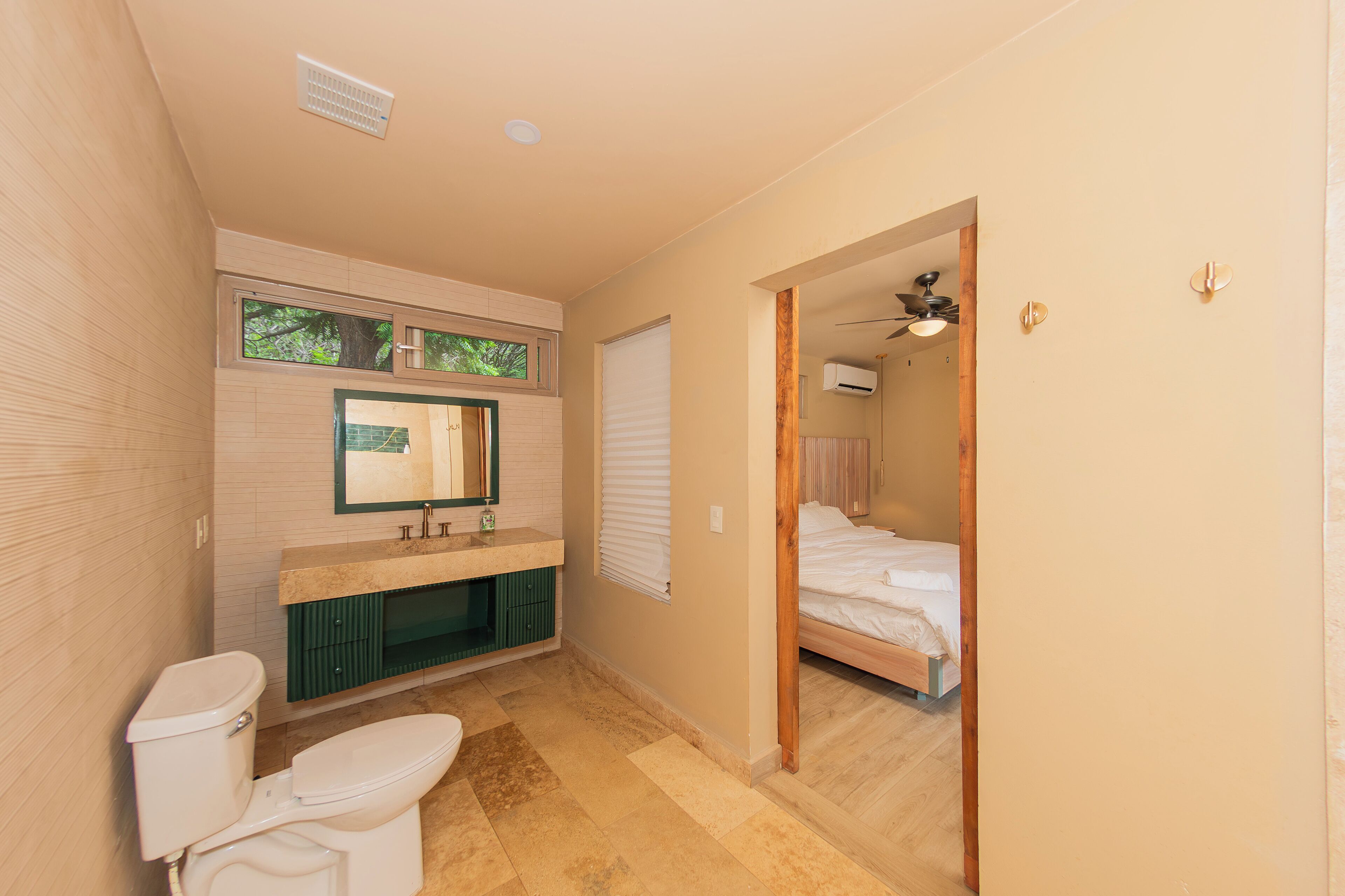 A bathroom with a toilet, sink, and mirror. There is a window with blinds and a door leading to a bedroom.