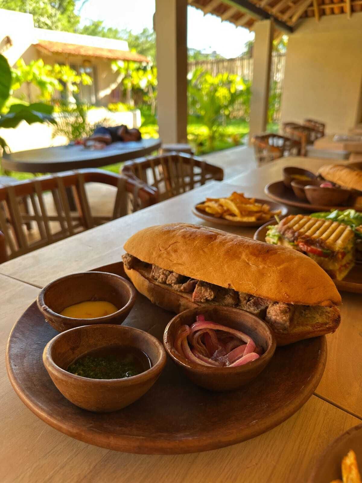 Un tavolo da pranzo con un grande panino, contorni e un vassoio di legno.