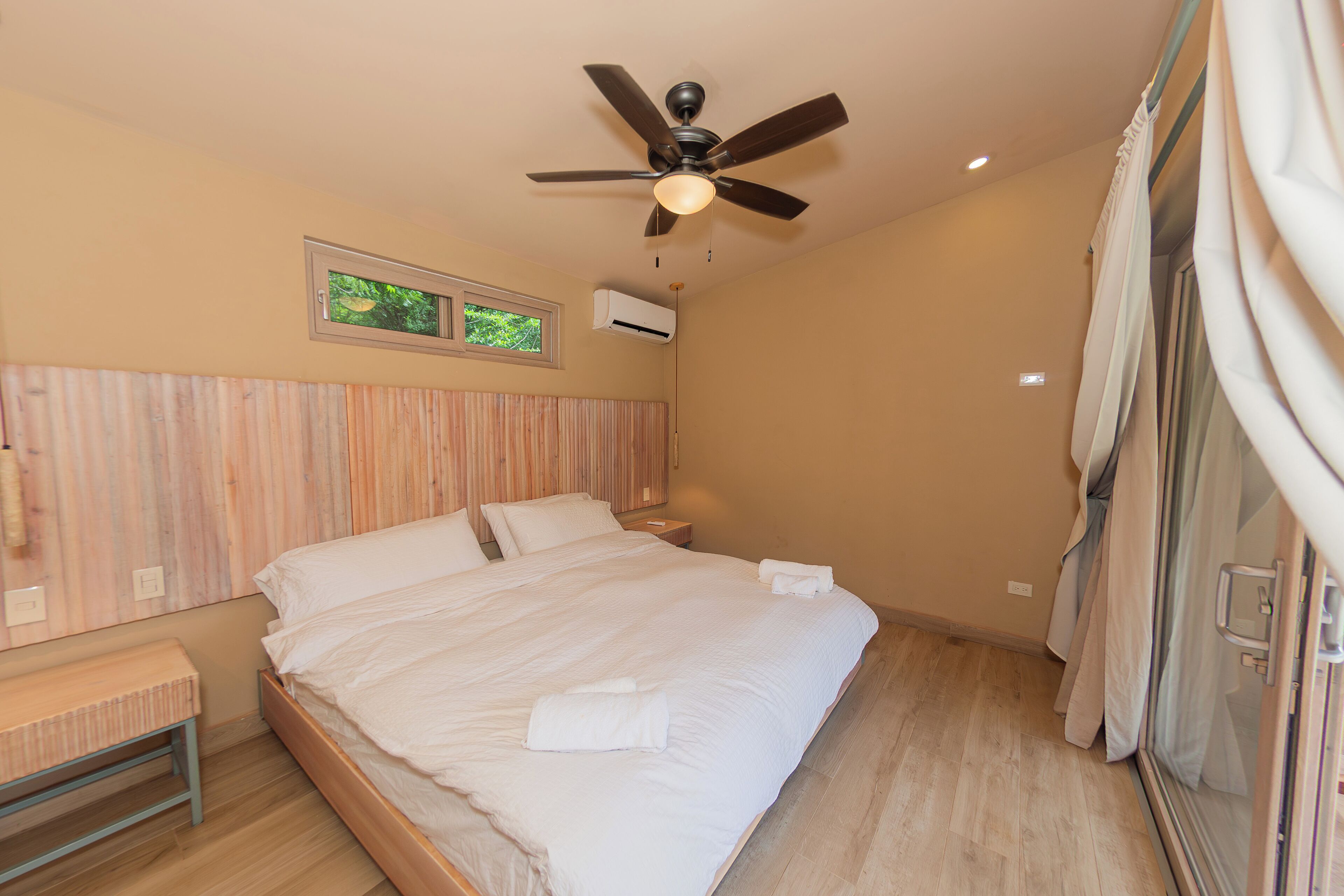 Un dormitorio con una cama, ventilador de techo, ventana y mesita de noche.