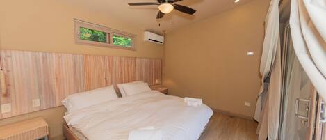 Un dormitorio con una cama, ventilador de techo, ventana y mesita de noche.