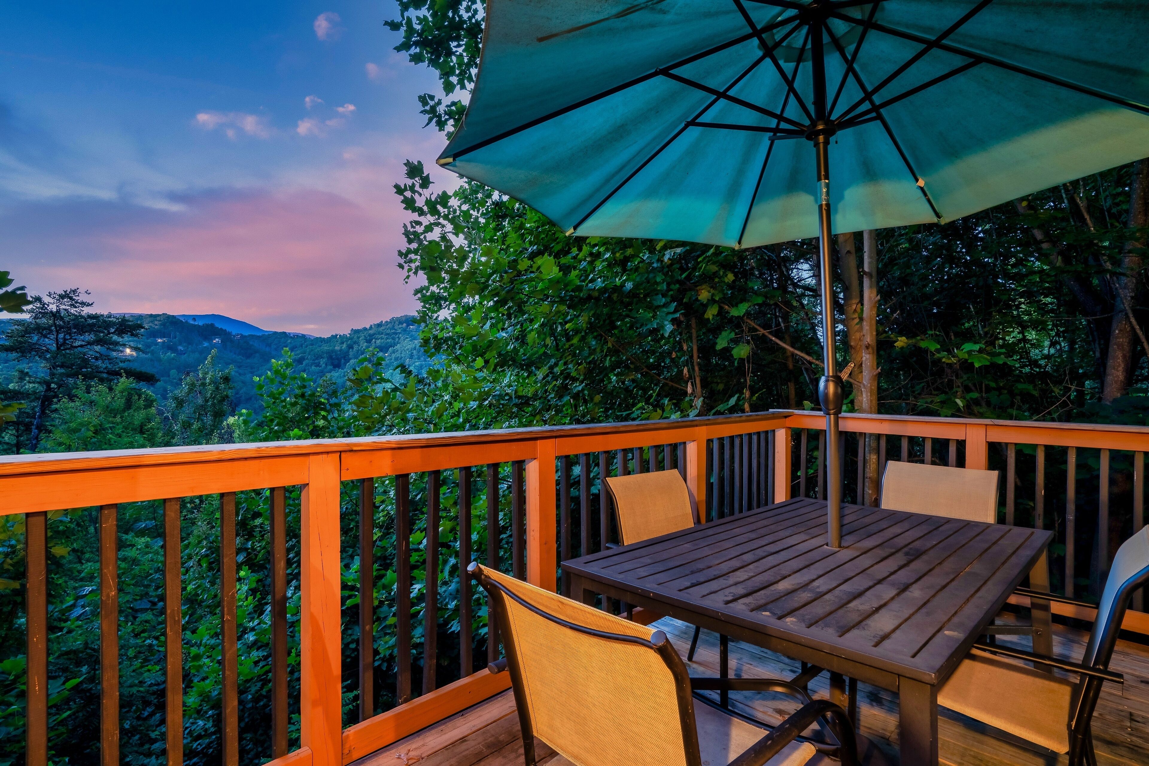 Um deck de madeira com mesa e cadeiras, rodeado por vegetação exuberante e vista para colinas ao longe.