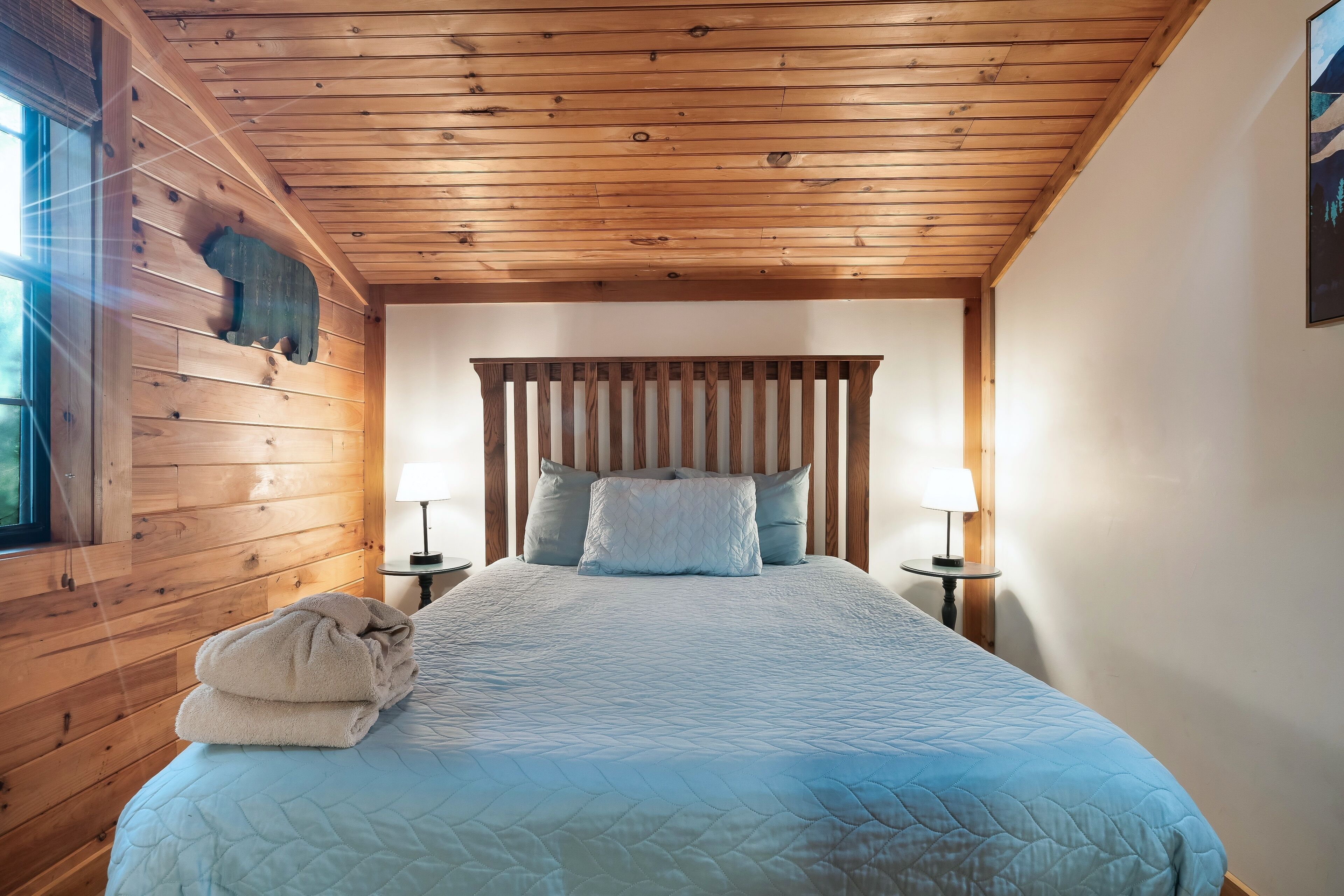 Un dormitorio con techo de madera, una cama con colcha azul acolchada, dos lámparas de noche y una ventana a la izquierda.