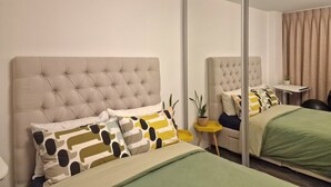 Ett sovrum med en stor spegel, två sängar med grönt och beige sänglinne och ett litet sidobord med en växt.