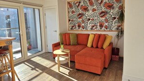 Ett modernt vardagsrum med en stor orange soffa, ett litet runt bord och en skjutdörr i glas som leder ut till en balkong.