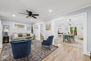 Ein geräumiges Wohnzimmer mit einer Couch, zwei blauen Sessel, einem Couchtisch und einem gemusterten Teppich. Eine Treppe führt in einen anderen Raum, und es gibt einen Essbereich mit Tisch und Stühlen.
