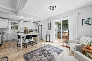Eine moderne Küche mit weißen Schränken, ein Essbereich mit einem weißen Tisch und schwarzen Stühlen sowie ein Wohnzimmer mit einem grauen Sofa und einem Holztisch.