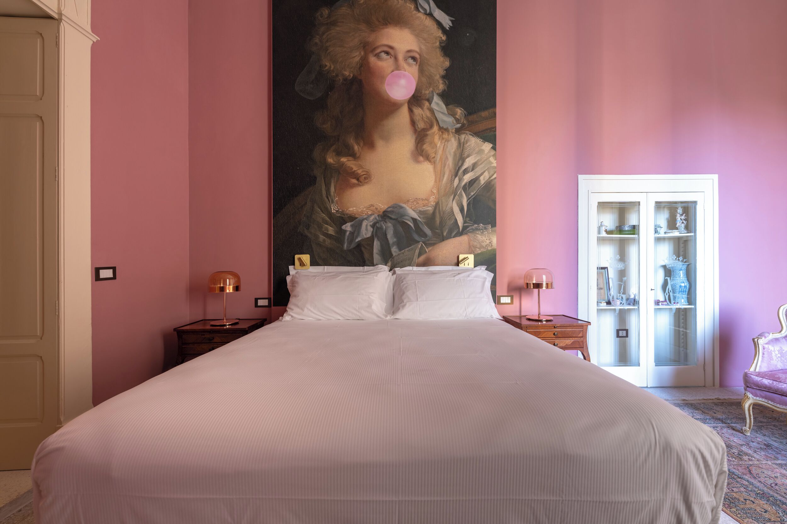 A large bed in a room with pink walls, a large painting, and a cabinet with glass doors.