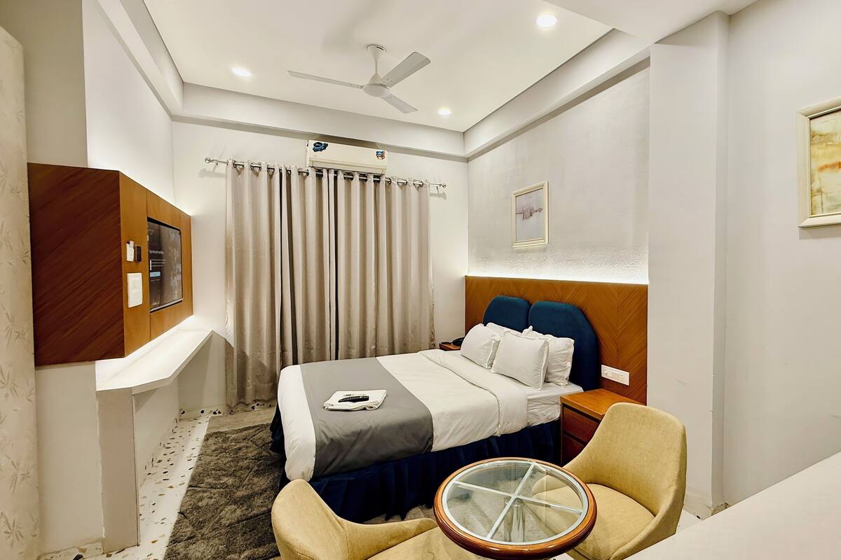 Habitación de hotel con cama, una silla, una mesita, ventilador de techo y ventana con cortinas.