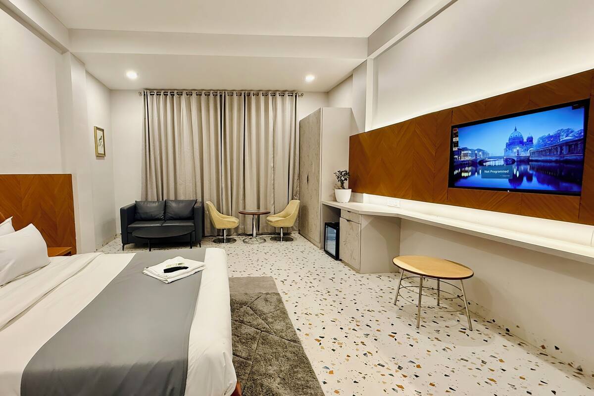 Habitación de hotel con cama, sofá, televisor montado en la pared, una mesita y una silla.