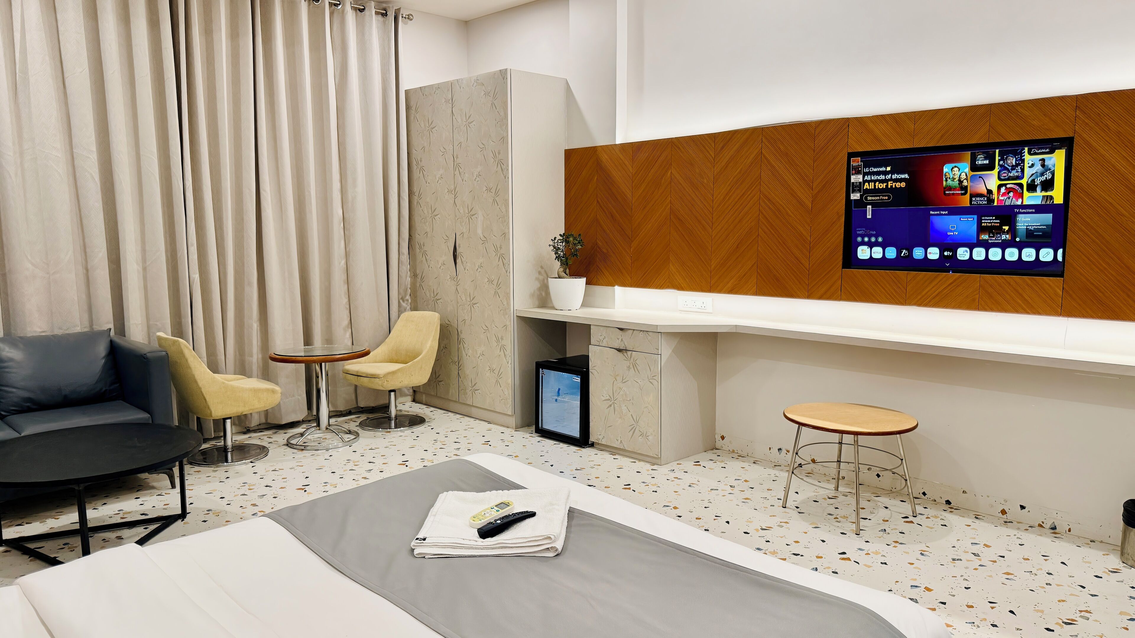 Een moderne hotelkamer met een bed, een bank, twee fauteuils, een bijzettafeltje, een televisie en een plant.