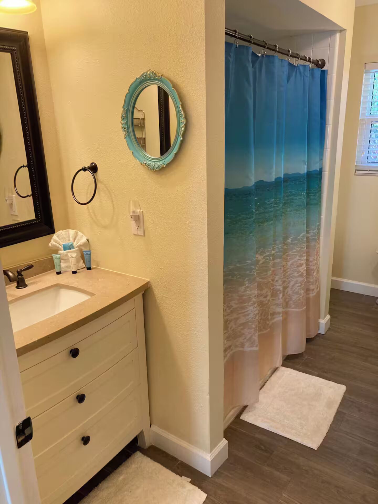 Un baño con tocador color beige, un espejo, un aro para toallas y cortina de ducha con una escena de playa.