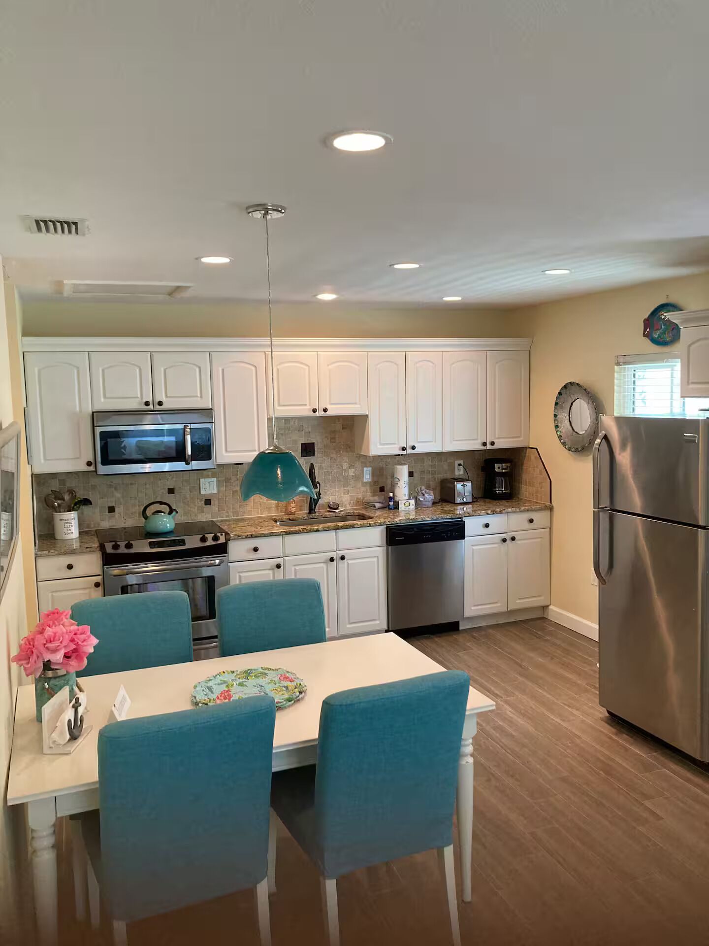 A modern kitchen with white cabinets, stainless steel appliances, a dining table with blue chairs, and a teal pendant light.