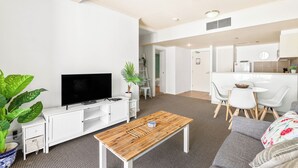 Una sala moderna con un televisor de pantalla plana, una mesa de centro de madera, un sofá gris y un comedor con una mesa y sillas blancas.