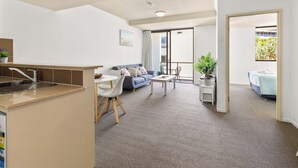 Una sala moderna con sofá, mesa de centro y zona de comedor con sillas. Incluye una cocina con pileta y mostrador. El piso está alfombrado y hay ventanas amplias.