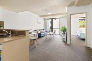 A modern living room with a sofa, coffee table, and a dining area with chairs. There is a kitchen area with a sink and a countertop. The room has a carpeted floor and large windows. - Burleigh Bliss Coastal Hideaway (Burleigh Heads)