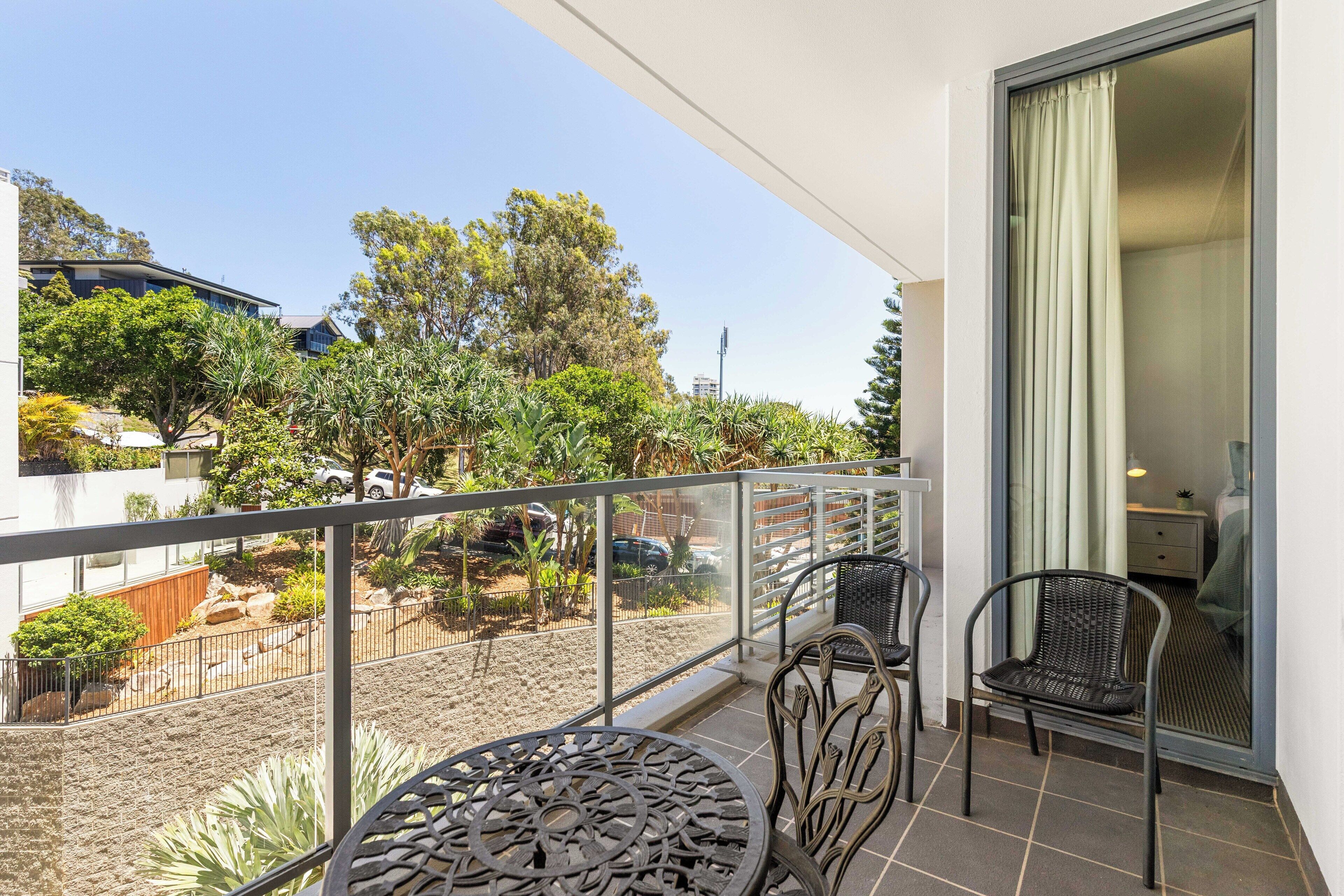 A balcony with a table and two chairs, overlooking a garden with various plants and a distant building.