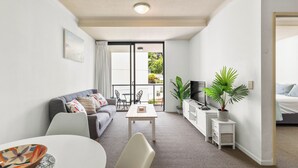 Una habitación de hotel moderna con sofá, mesa de comedor, televisor, planta en maceta y balcón con vistas.