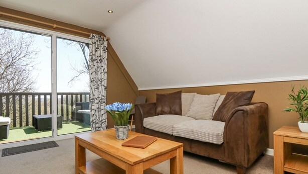 A cozy living room with a brown sofa, a wooden coffee table, and a view of a balcony with a wooden railing. - Valley Lodge 61 (Gunnislake)