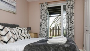 A bedroom with a bed, a nightstand, a lamp, and a view of the outside through a window with patterned curtains. - Valley Lodge 61 (Gunnislake)