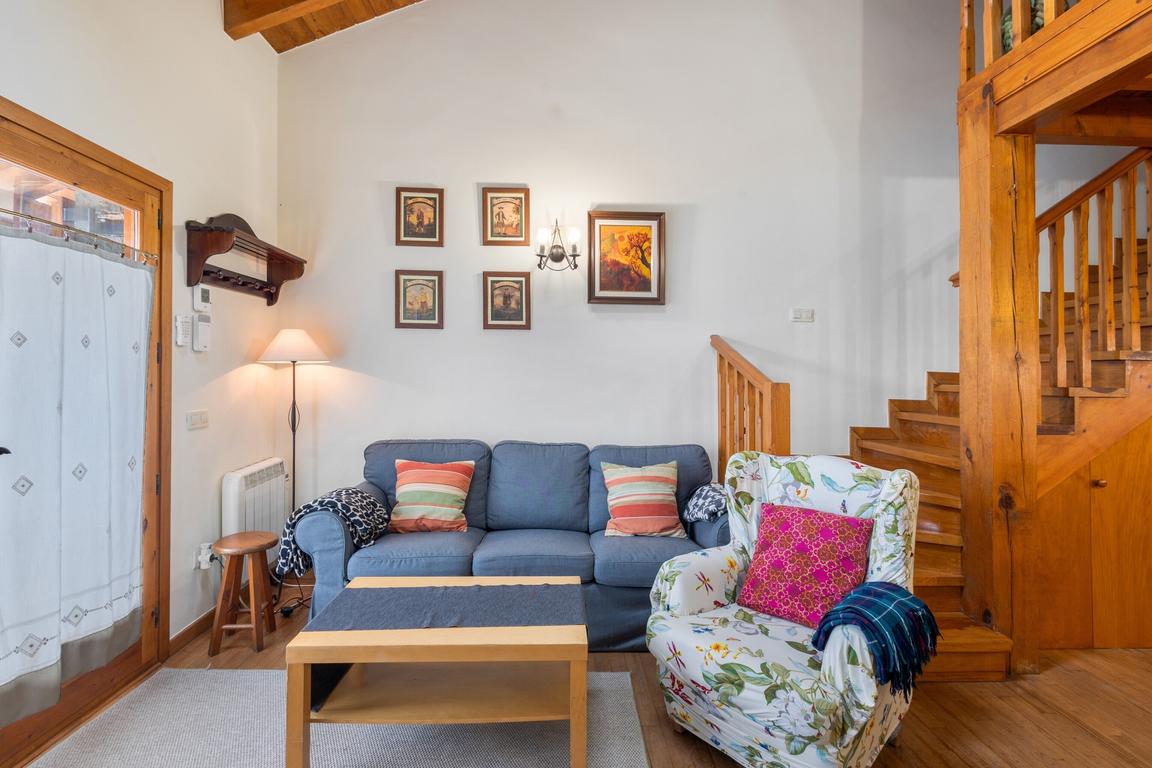 Een woonkamer met een bank, fauteuil, houten trap en aan de muur hangende foto’s.