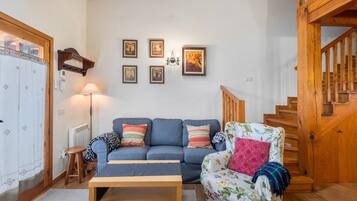 Een woonkamer met een bank, fauteuil, houten trap en aan de muur hangende foto’s.