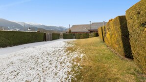 Un sendero cubierto de nieve que lleva a un edificio con techo tradicional, rodeado de setos recortados y montañas al fondo.