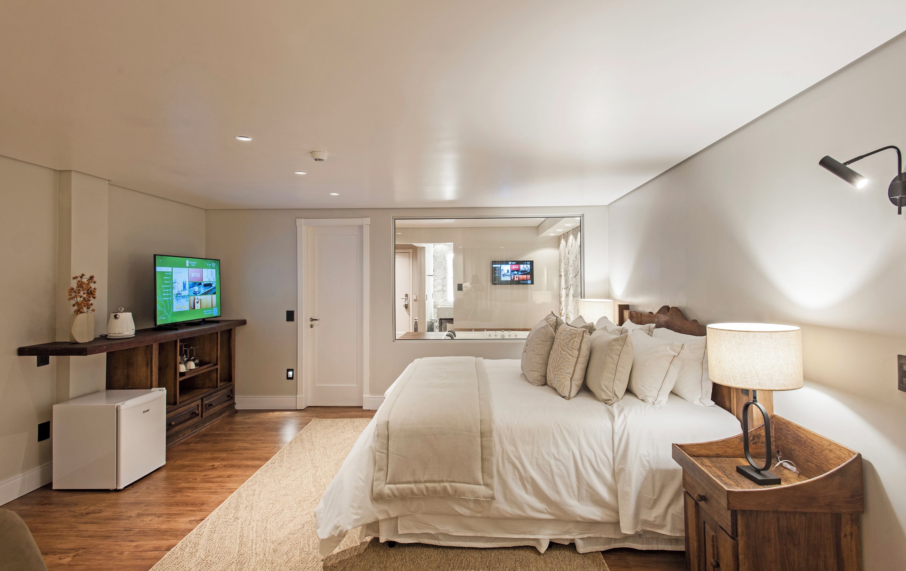 A modern bedroom with a bed, a TV, a small refrigerator, and a bedside table with a lamp.