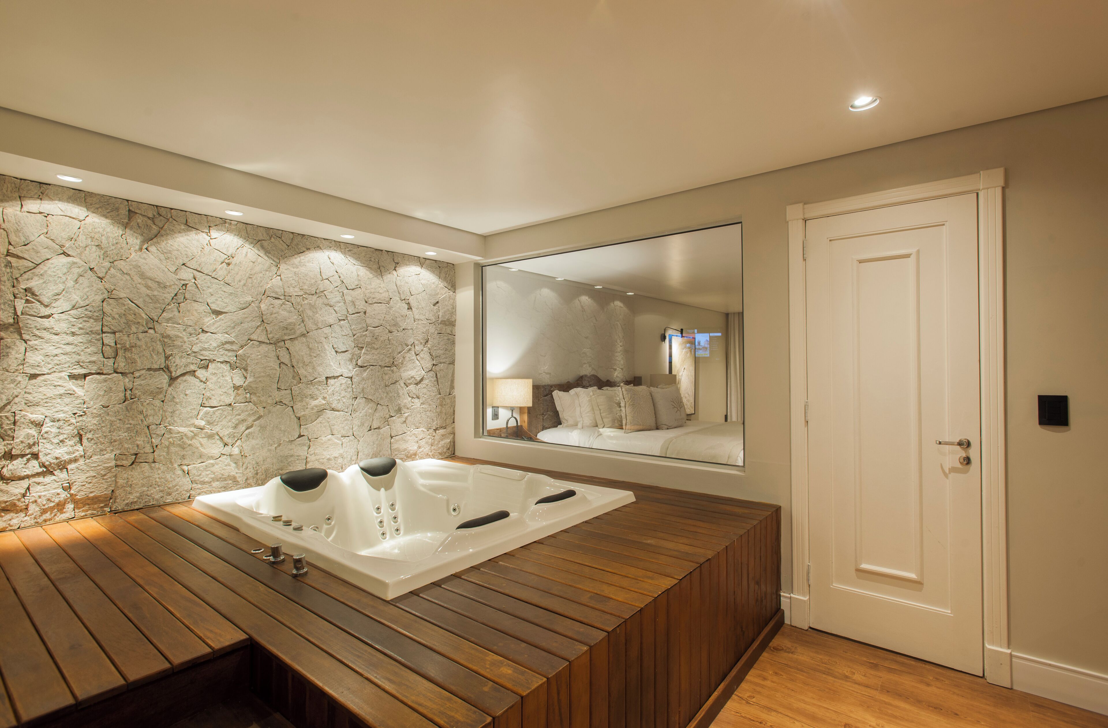 A modern bathroom with a stone wall, a wooden bench, a large bathtub, and a mirror reflecting a bedroom with a bed and a nightstand.