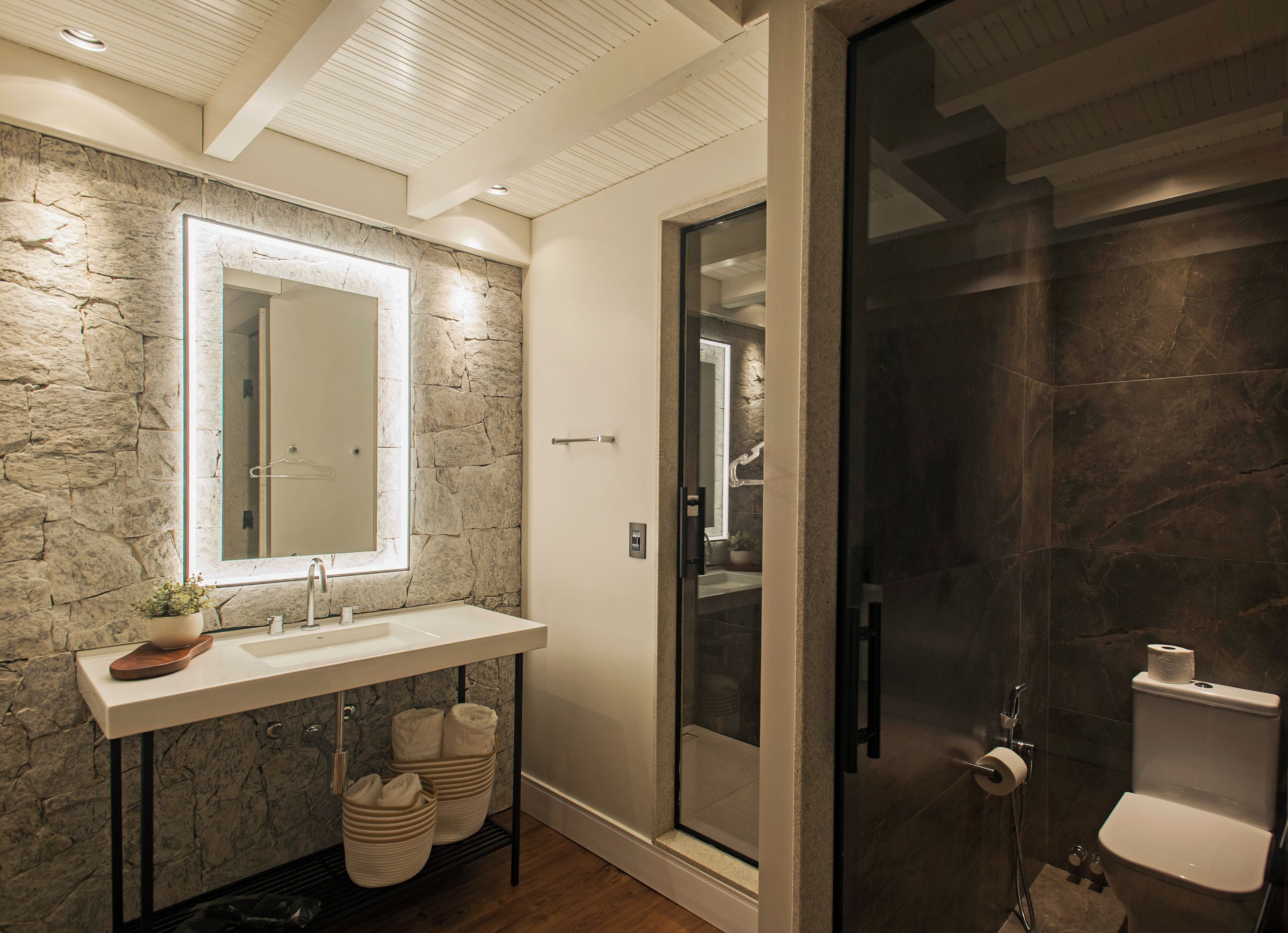 Un baño moderno con pared de piedra, un espejo grande, un lavabo blanco y zona de ducha con mampara de cristal.