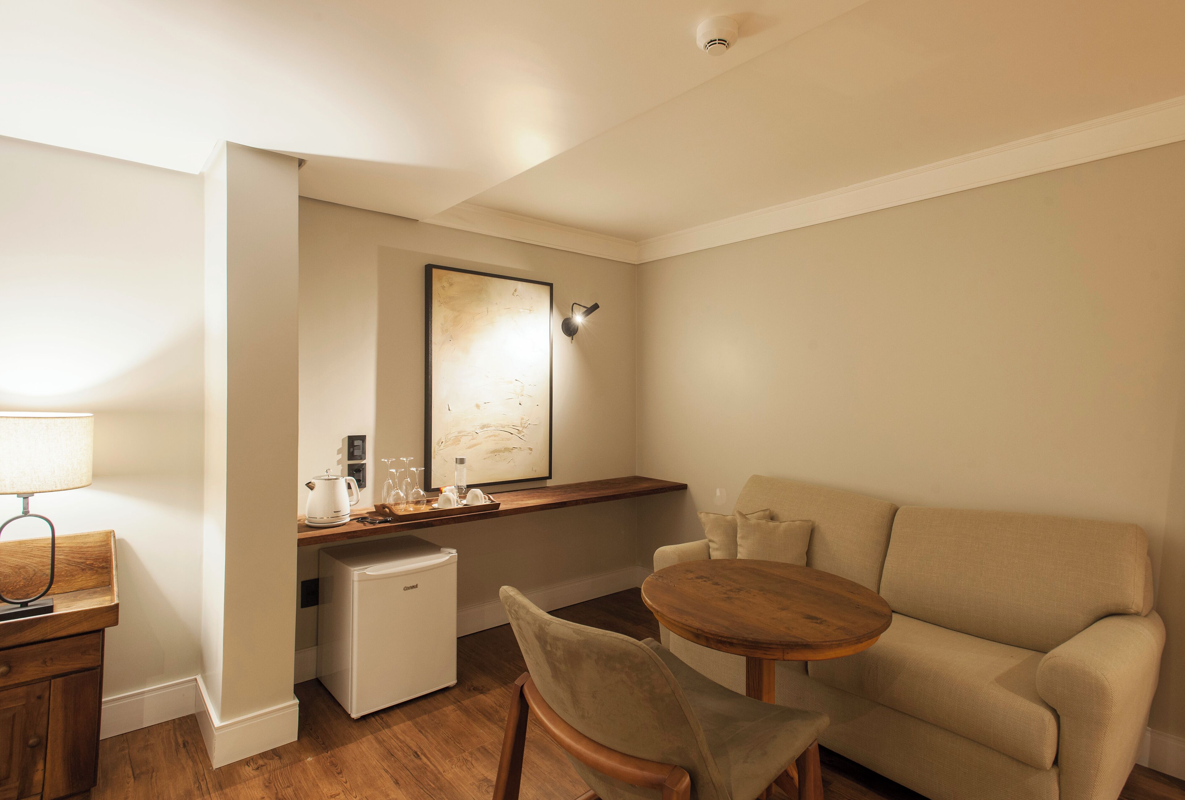 A compact living space with a kitchenette, a small refrigerator, a sofa, a wooden table, and a lamp.