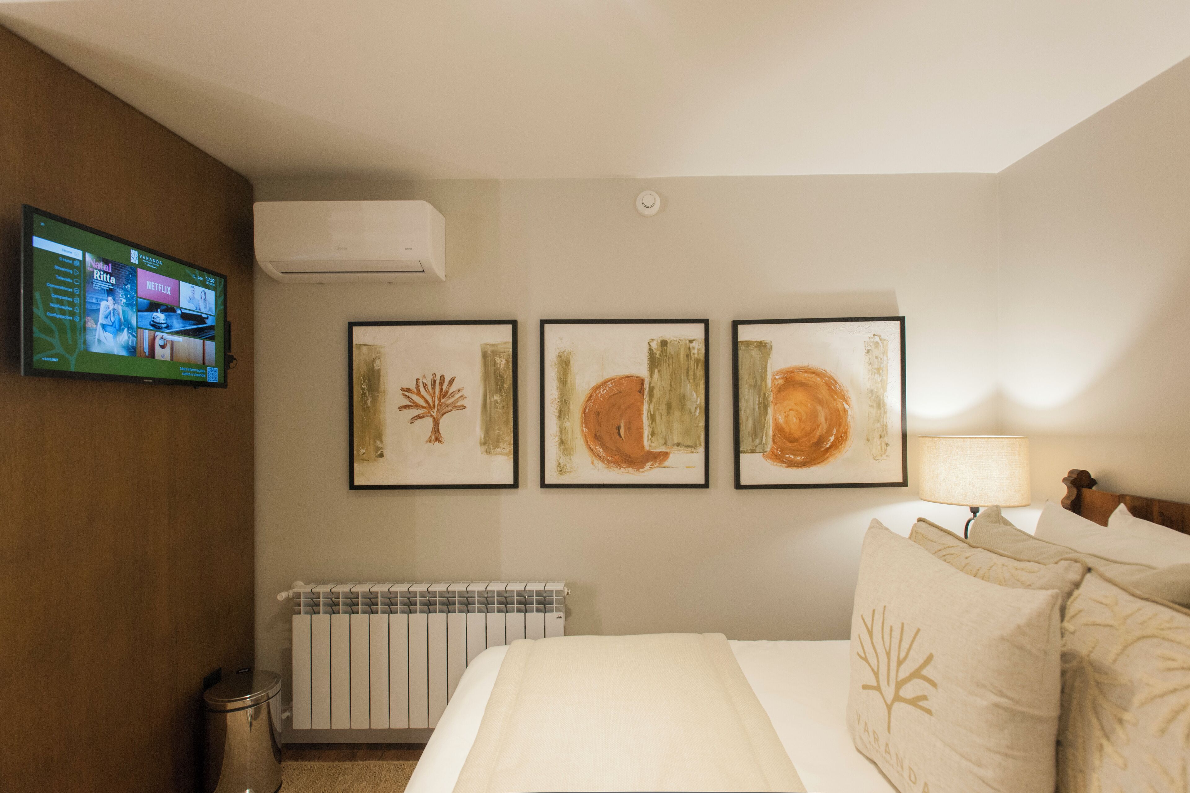 A hotel room with a bed, a TV mounted on the wall, and three framed artworks above the bed.