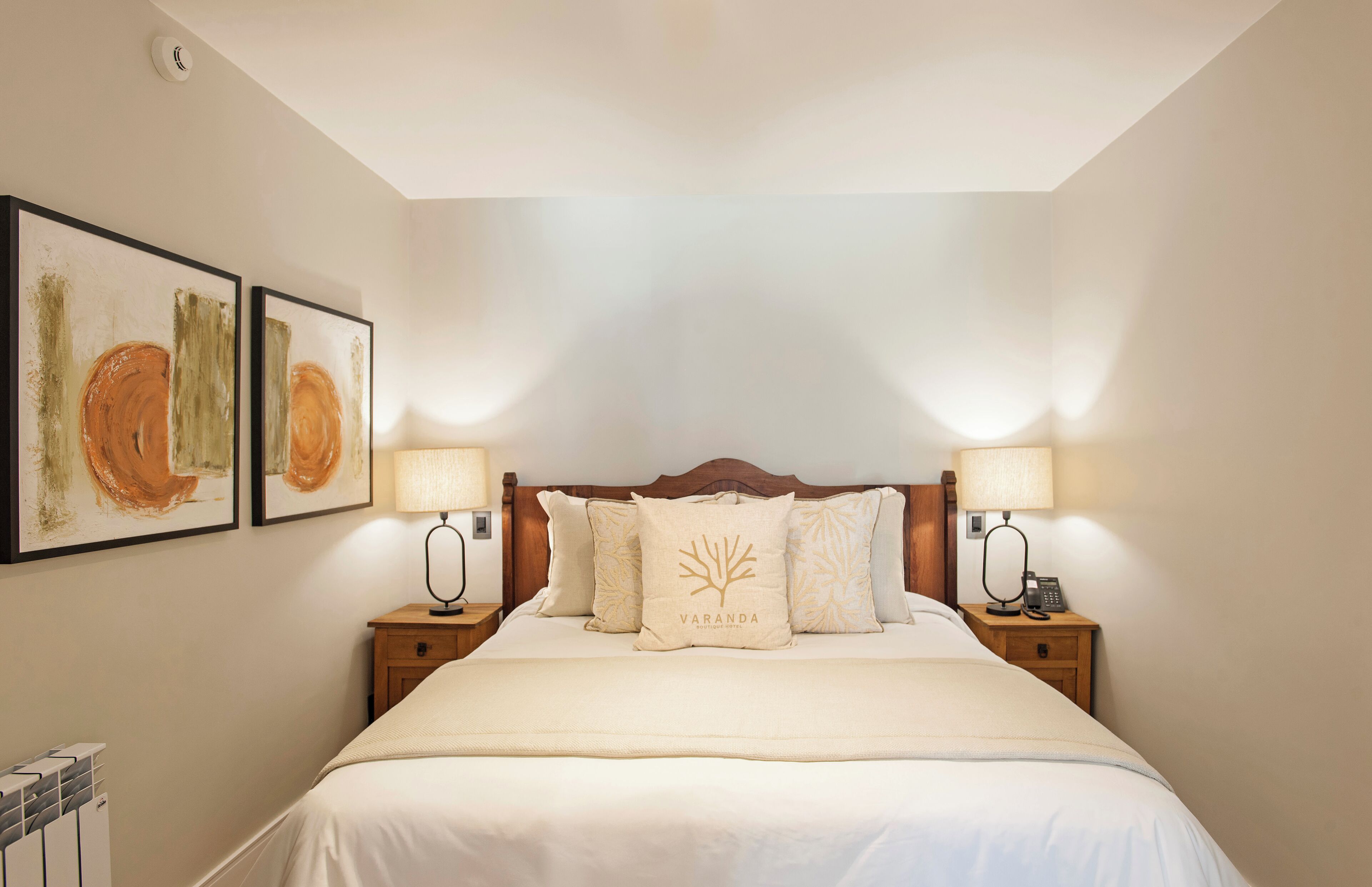 Une chambre d’hôtel bien rangée, avec un grand lit, deux tables de chevet avec des lampes et deux tableaux abstraux encadrés accrochés au mur.