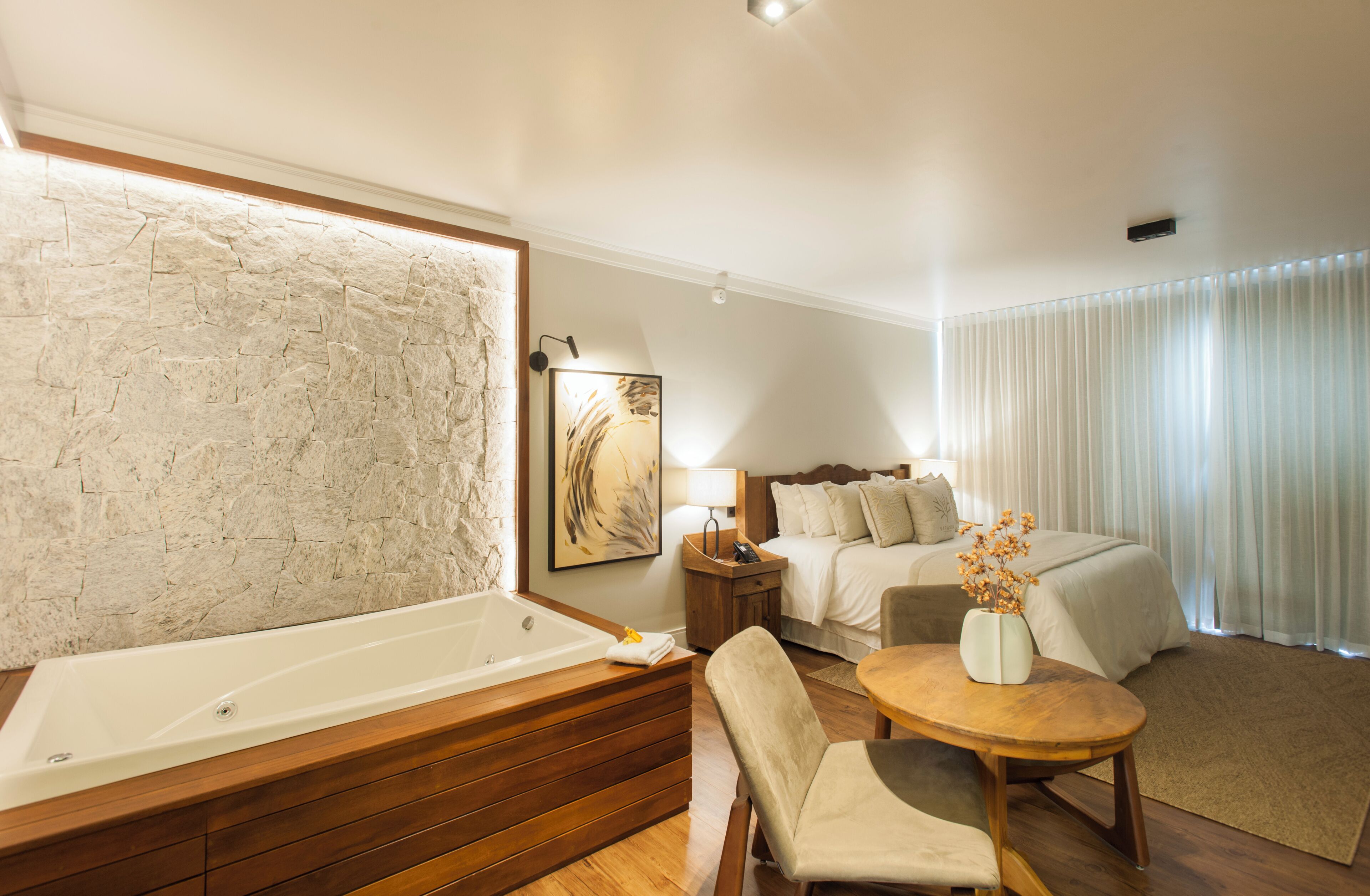 A modern hotel room with a large bathtub, a bed with multiple pillows, a bedside table with a lamp, and a small round table with a vase of flowers.