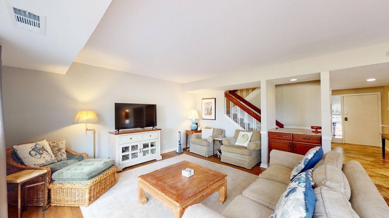 Een ruime woonkamer met een grote bank, een houten salontafel en een televisie. Er is een trap die leidt naar een ander niveau, en een witte kast met lades.